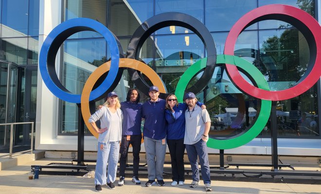 Five people standing and smiling in front of the Olympic Rings, which are large, colored circles.