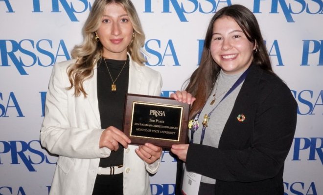 Two college students dressed professionally standing in front of a white backdrop with blue lettering. They are holding an award.