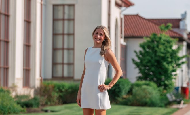 A young woman in a white dress standing proudly on a college campus with a white building in the background.
