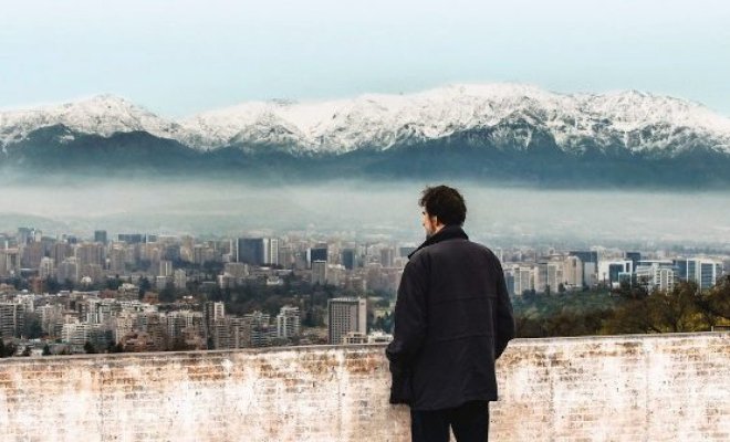 A man stands at a wall and looks over a cityscape with a large mountain range in the background