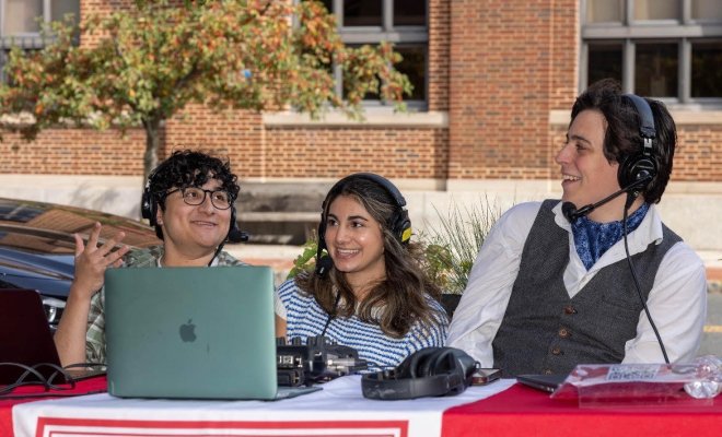 Three young students sitting outside at a table with a laptop in front of them. All of them are wearing headphones and are part of a live radio broadcast. They are smiling and happy.