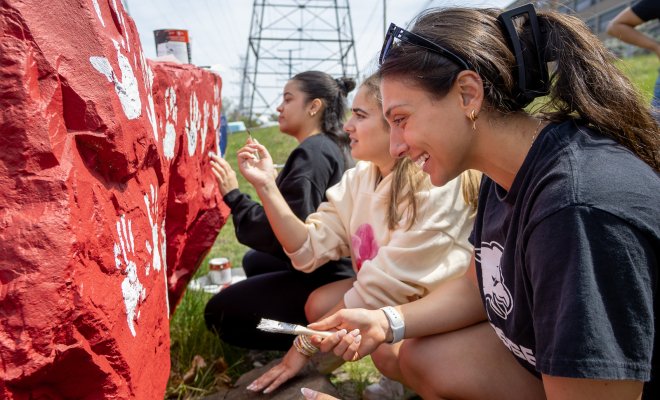 Three students smile as they paint white handprints onto a large red campus rock during an outdoor community service activity.