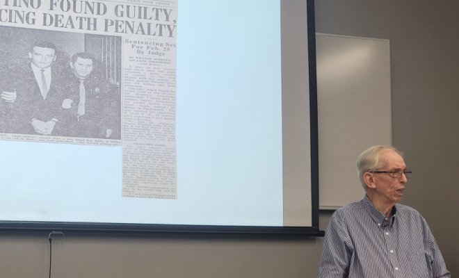 An old man stands in front of a projector screen and delivers a lecture
