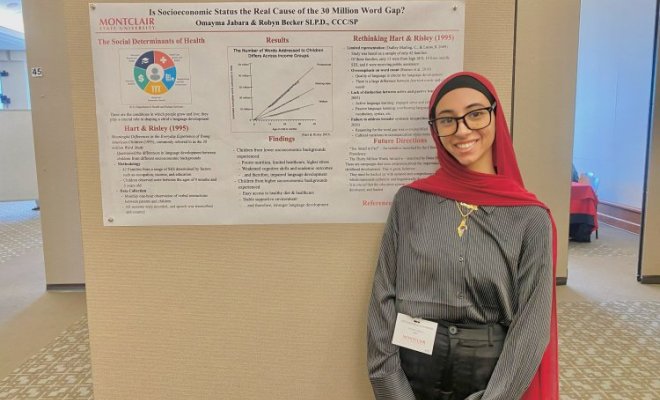A college student wearing a black head covering and a long red scarf stands in front of a printed presentation pinned to a wall
