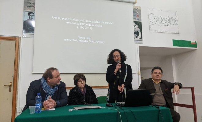Four people stand at a presentation table and one stands and holds a microphone