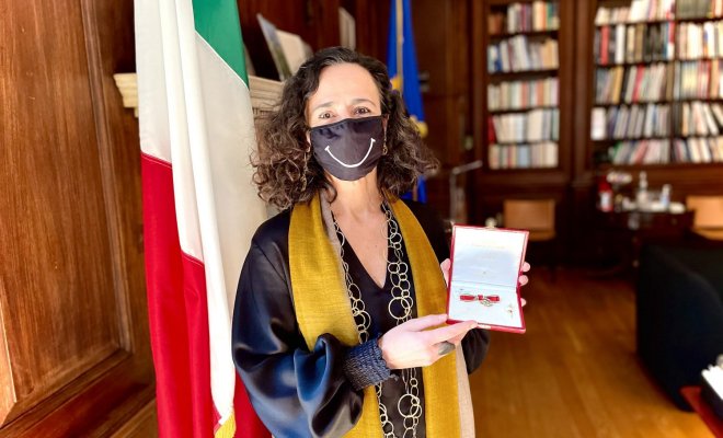 A woman stands in front of an Italian flag and holds open a display box 