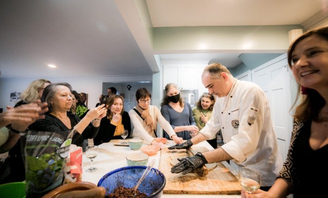 A chef rolls dough on a table while a group of people watch and take photos