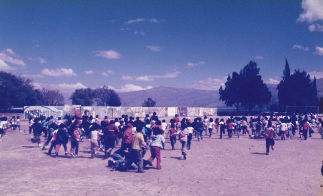 A large grouup of children of various ages run around an outdoor dirt field