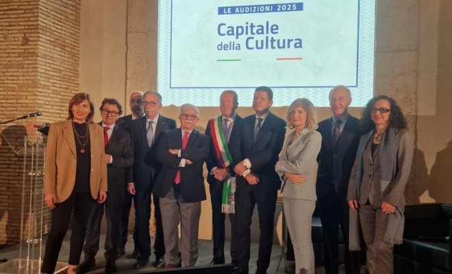A group of people pose in front of a projector screen bearing the words "Le Audizioni 2025 Capitale della Cuultura""