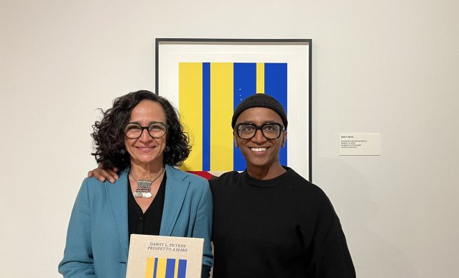 Two people pose in front of framed art piece in a gallery