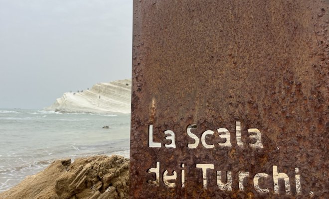 White cliffs along the ocean with a rusted piece of  metal with the words "La Scala dei Turchi" in the foreground