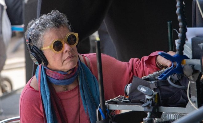 A woman wears sunglasses and headphones and looks toward a screen