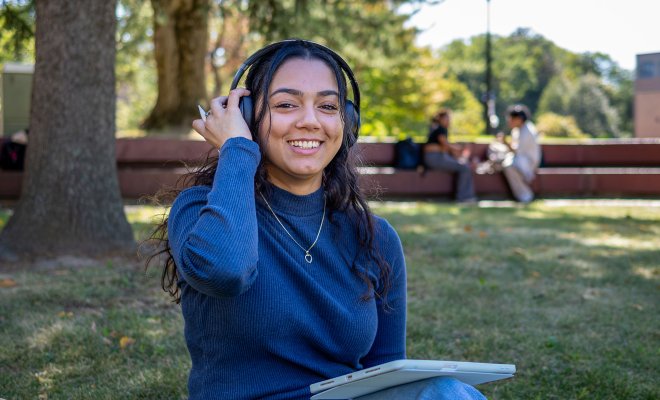 Student wearing headphones and smiling at the camera. They have an ipad on their lap.