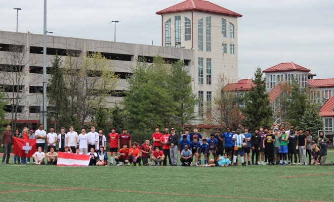 A large group of college students in soccer attire stand along the edge of a pitch and hold flags