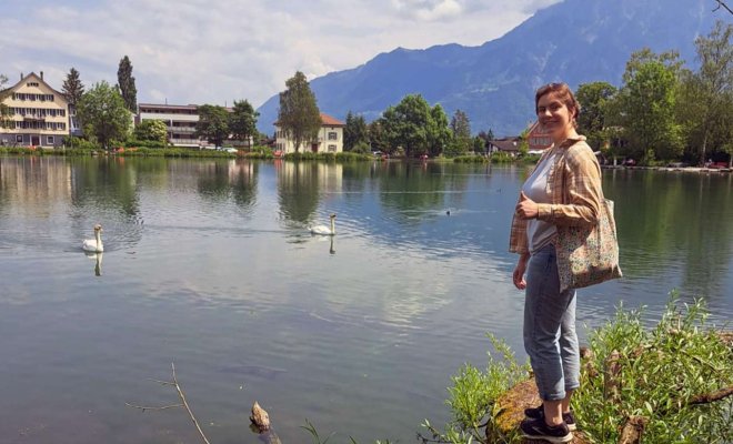 A woman stands at the edge of a lake with mountains and buildings across from her
