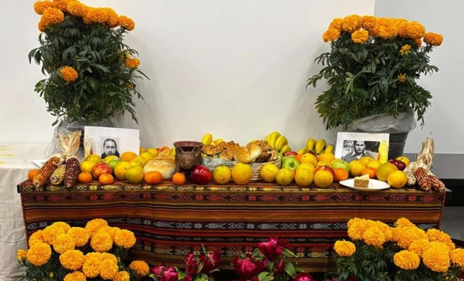 Día de los Muertos altar decorated with orange marigolds, fruit, bread, corn, photographs, and other offerings arranged on a colorful woven cloth