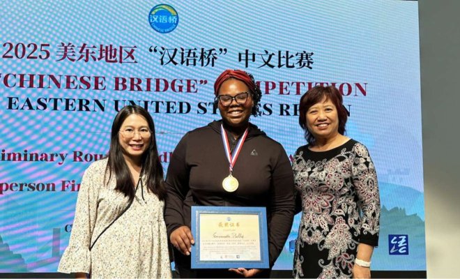 Three women pose for a photo in front of a projection screen, the woman in the middle wears a medal and holds a framed certificate