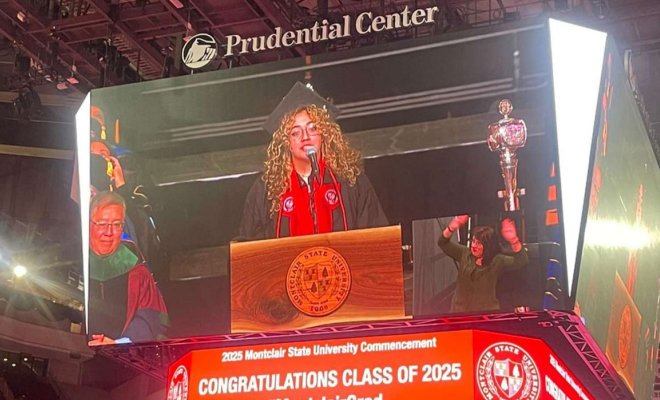 On a video board, a woman wearing graduation regalia speaks at a podium