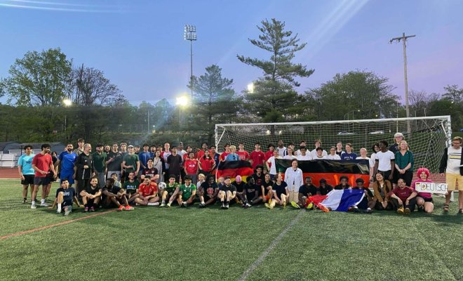 A group of college students and professors hold flags and pose in front of a goal on a soccer field
