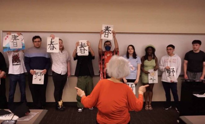 A group of college students stand in a line holding papers with Japanese vocabulary as they sing and follow an instructor's lead