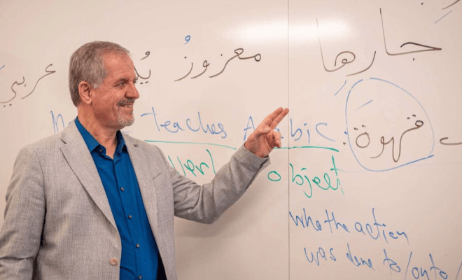 A man in a suit stands in front of a whiteboard and points to a piece of writing