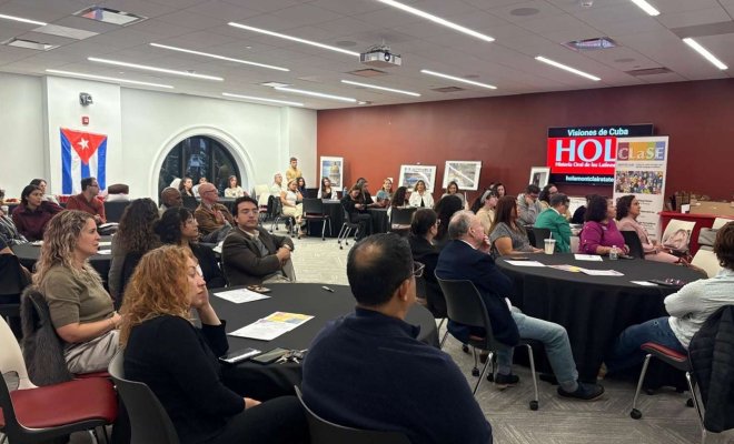 People sit around several round tables in a conference room with a video board reading "Visiones de Cuba" and watch a presenter 