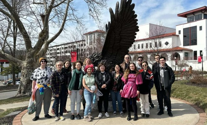 A group of college students pose together in front of a metal statue of a Red Hawk