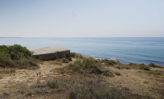 Dry coastal bluff with scrub vegetation and a low, defunct World War II concrete bunker overlooking a calm blue sea under a hazy sky