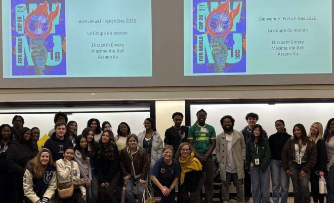 A large group of college students pose in front of two project screens in a classroom
