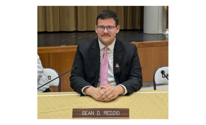 A man wears a suit and tie and sits at a desk with a name plate in front of it