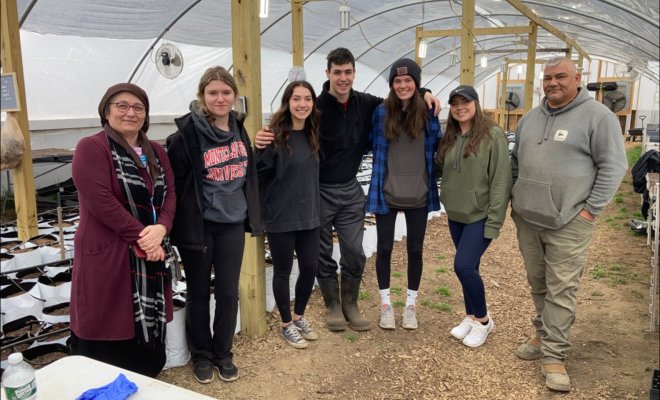 A group of people wearing cold weather attire stand in side a greenhouse and pose