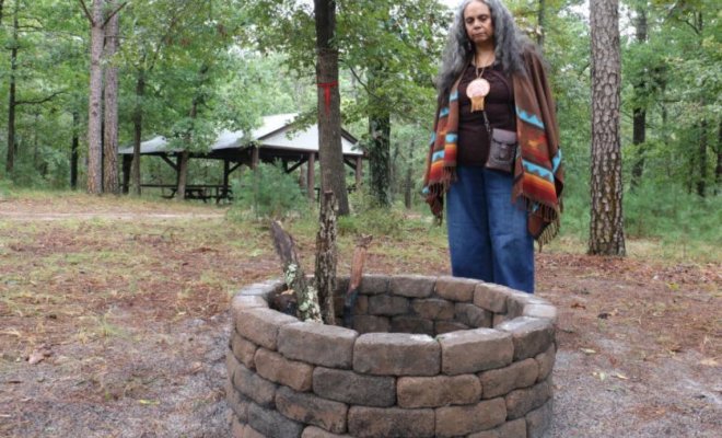A woman stands beside a circular stone-lined well in a wooded area, looking down into it. She wears a multicolored shawl and pendant, with a picnic shelter visible among the trees in the background