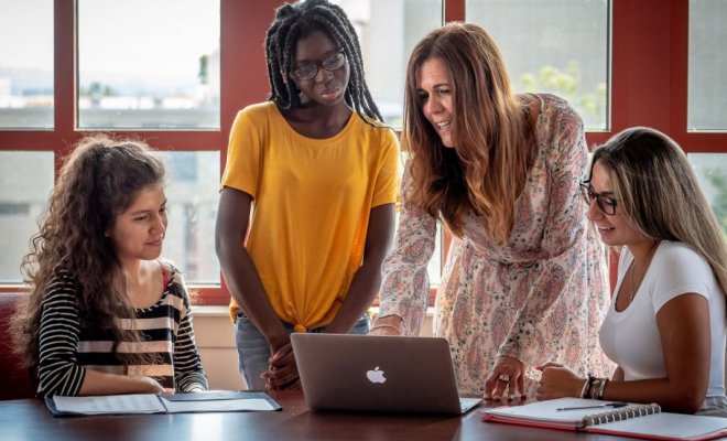 A college professor stands over a laptop and types while three students watch