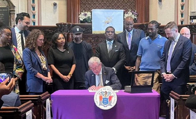 A group of people in professional clothing stand around a man seated at a table who is signing a paper