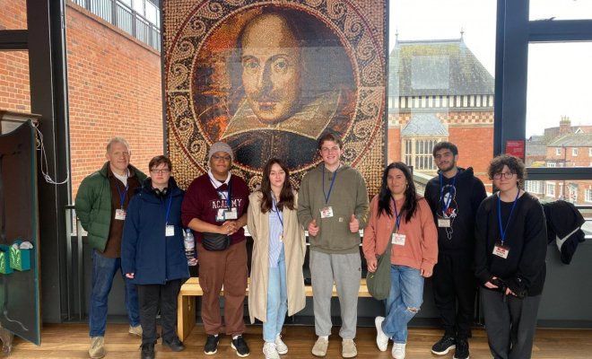 A group of people pose indoors in front of a tapestry of William Shakespeare's face.
