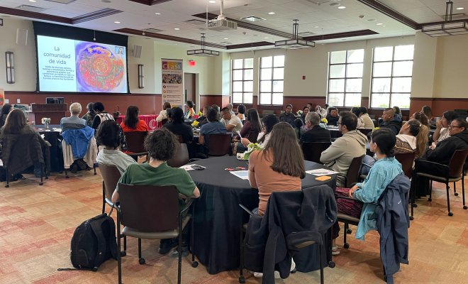 A crowd of people sit at round tables in a conference room and watch a projector screen