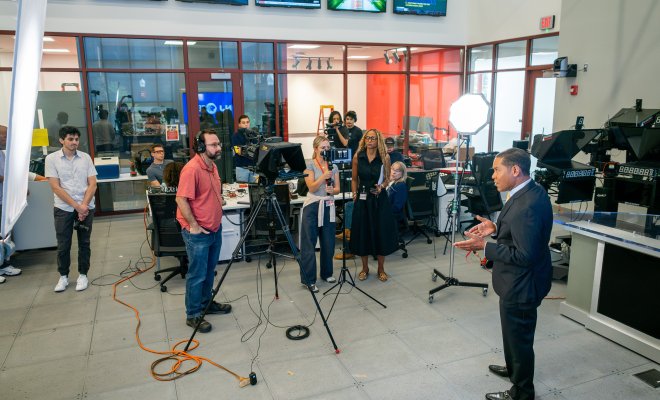 Rob Nelson, anchor of “NJ PBS News with Rob Nelson,” talks to a camera surrounded by faculty and students in newsroom. 