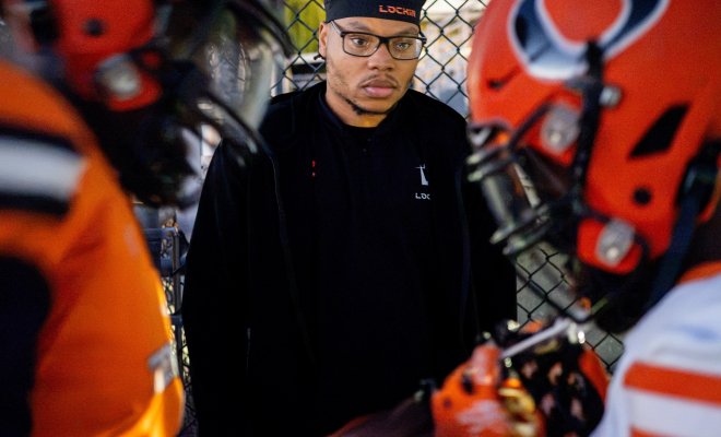 Montclair alumnus Emmanuel Ozias ’25, wearing a black Lock1n cap and jacket, stands by a chain-link fence surrounded by Orange High School football players in orange helmets and uniforms.