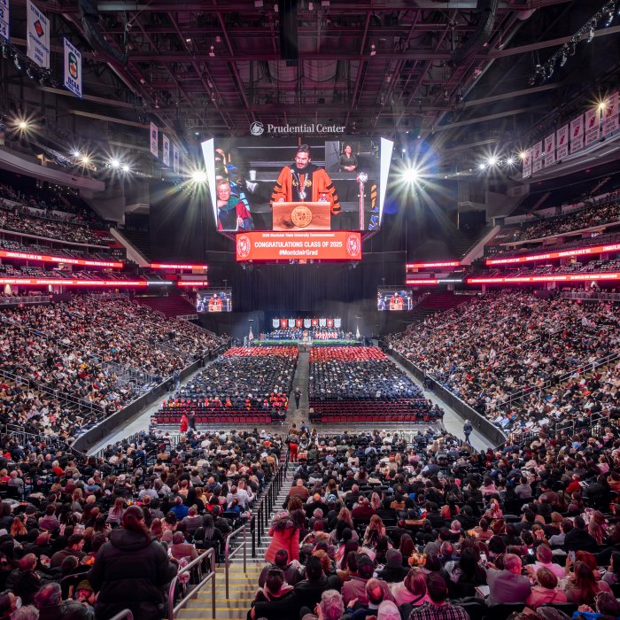 A wide shot of the Prudential Center captures the large crowd in the arena, with graduates seated on the floor in their caps and gowns. Above them, on the jumbo screen, the University president is visible, addressing the audience.