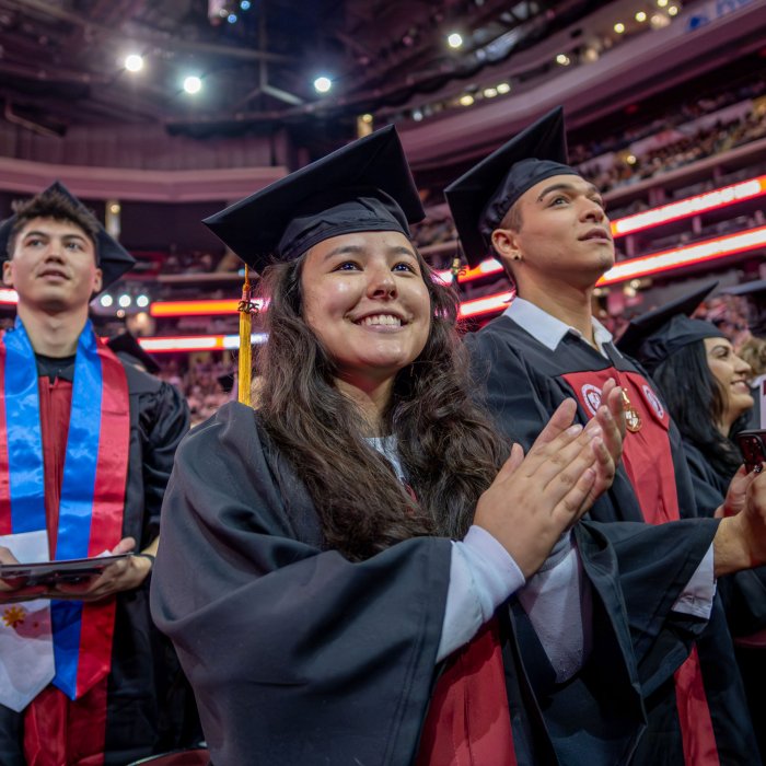 Three graduates raise their sights into the arena. In the center, one graduate smiles and claps,