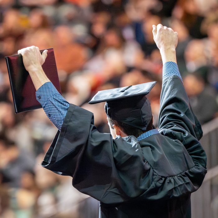A graduate raises his arms in triumph after receiving his diploma, shown from the back.