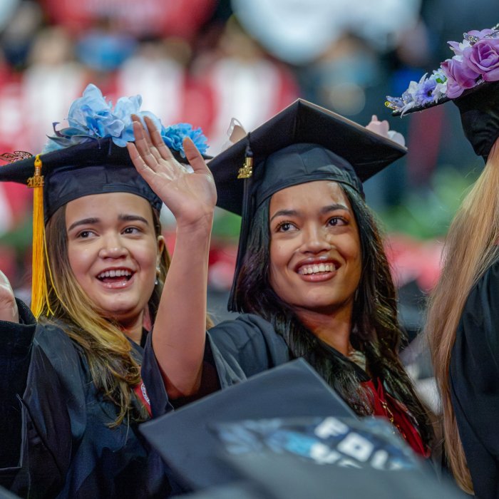 Two graduates, seated on the floor of the Prudential Center, turn and wave. Their caps are decorated with flowers.