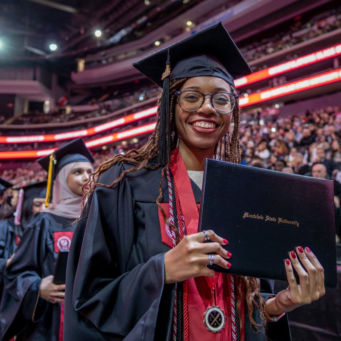 A graduate in a black gown and wearing glasses proudly holds up her diploma.
