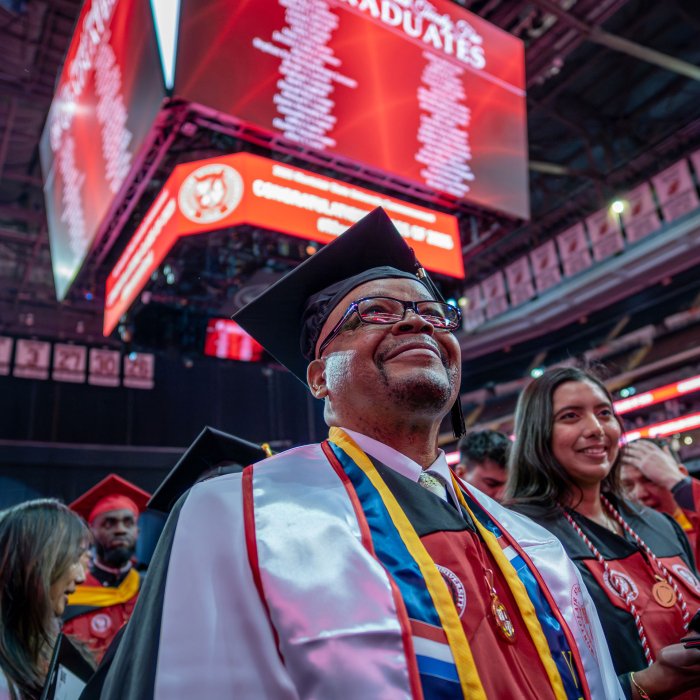 A graduate in a white stole looks up as names of all the graduates scroll across the jumbo screen above.