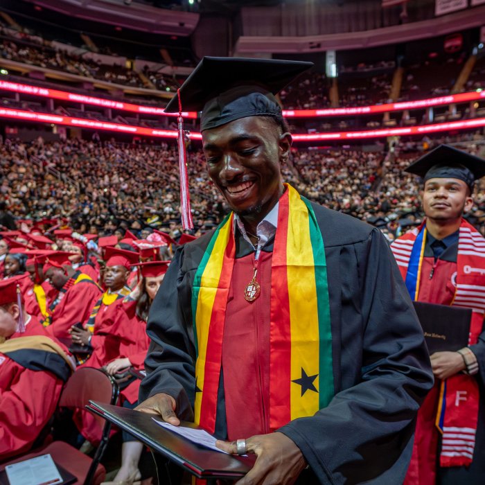 A graduate wearing a stole in red, yellow, and green with a black star proudly looks at their diploma after receiving it.