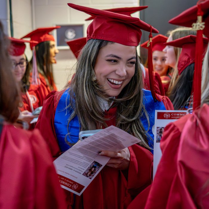 A graduate in a red gown smiles, surrounded by fellow graduates, while holding the day's program.