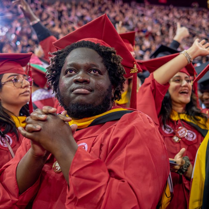 A graduate in a red gown clasps their hands, surrounded by fellow graduates in similar attire during the commencement ceremony.