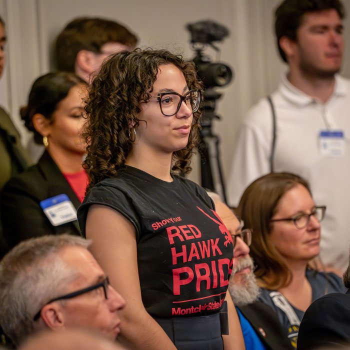 A woman in a Red Haw Pride t-shirt stands in the back of the hearing room.