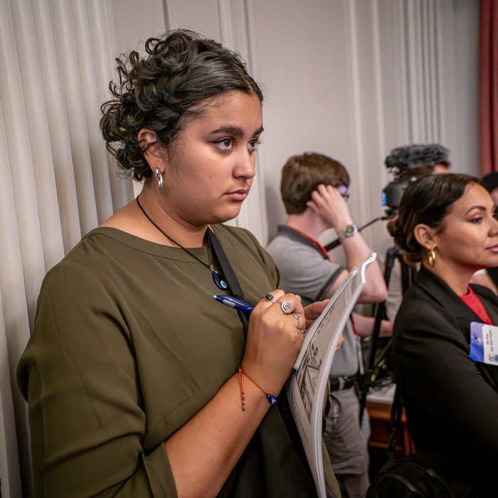 A woman standing in the back takes notes.
