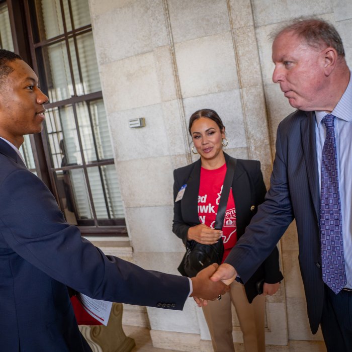 Two men shake hands as a woman in a Red Hawk Day t-shirt looks on.